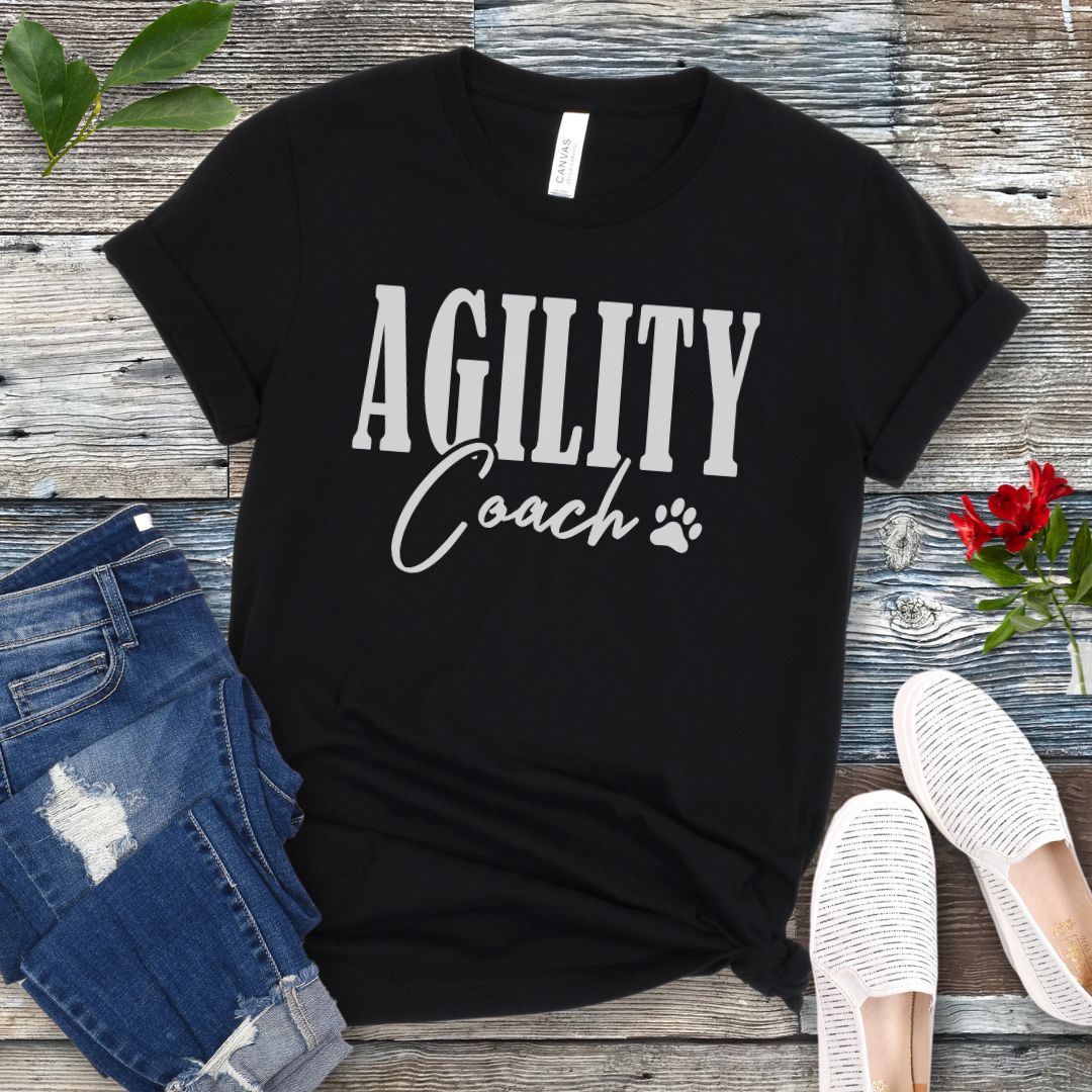 Black dog agility training t-shirt with 'AGILITY Coach' text in bold modern lettering, with accompanying paw print, shown on a wooden surface with jeans and shoes.