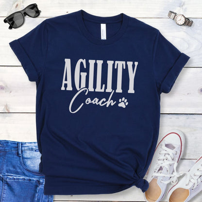 Black dog agility training t-shirt with 'AGILITY Coach' text in bold modern lettering, with accompanying paw print, shown on a wooden surface with jeans and shoes.