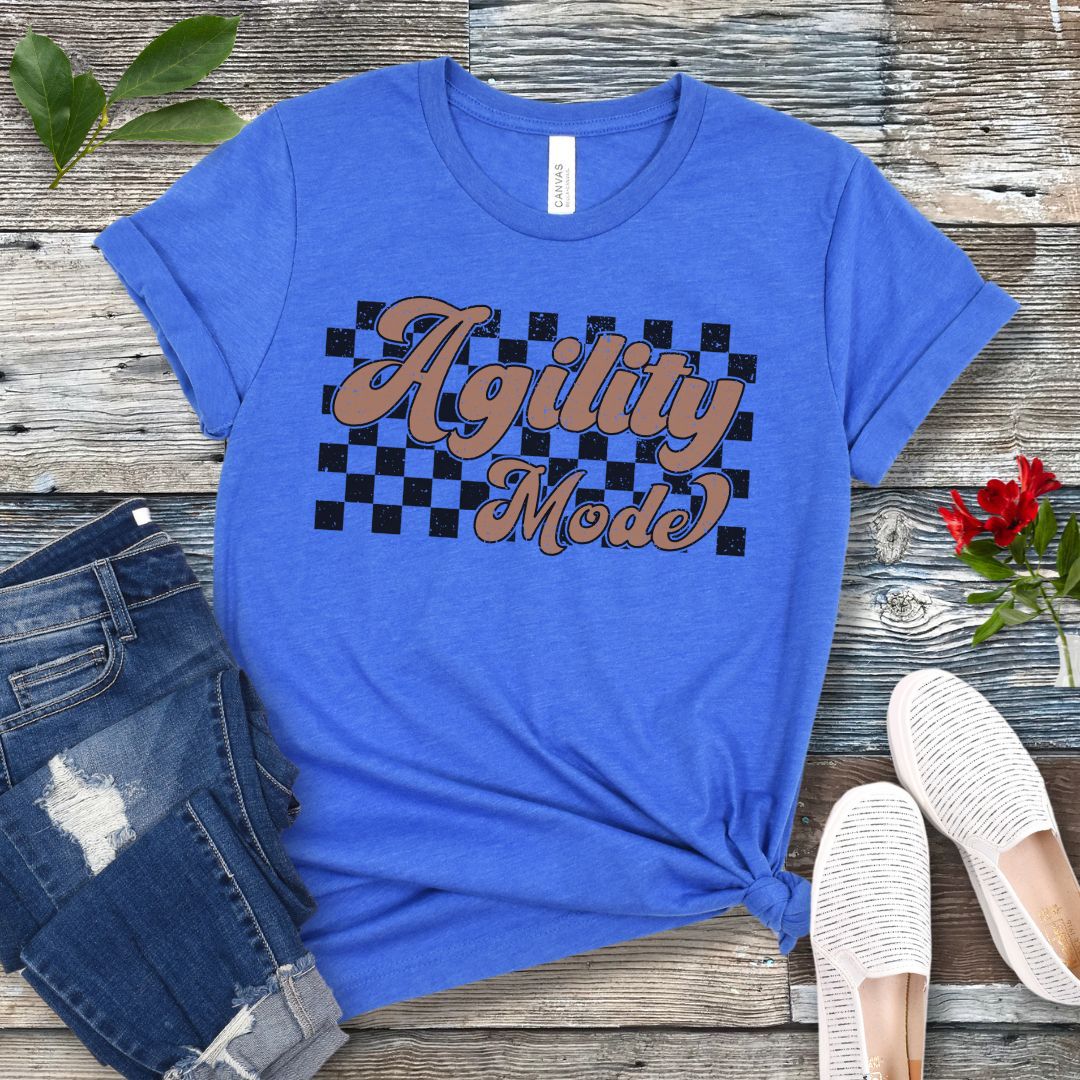 A blue dog agility t-shirt with 'Agility Mode' written in a retro stylized font over a checkerboard pattern, placed on a wooden surface, accompanied by jeans and white shoes.