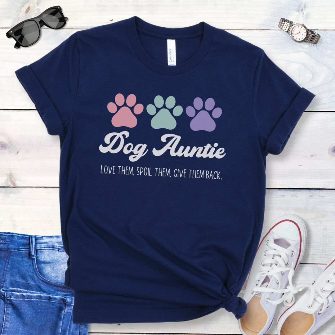 A navy blue dog lover t-shirt with 'Dog Auntie, Love Them Spoil Them, Give Theme Back' text and colorful paw prints design, placed on a wooden surface with jeans and white shoes in the background.