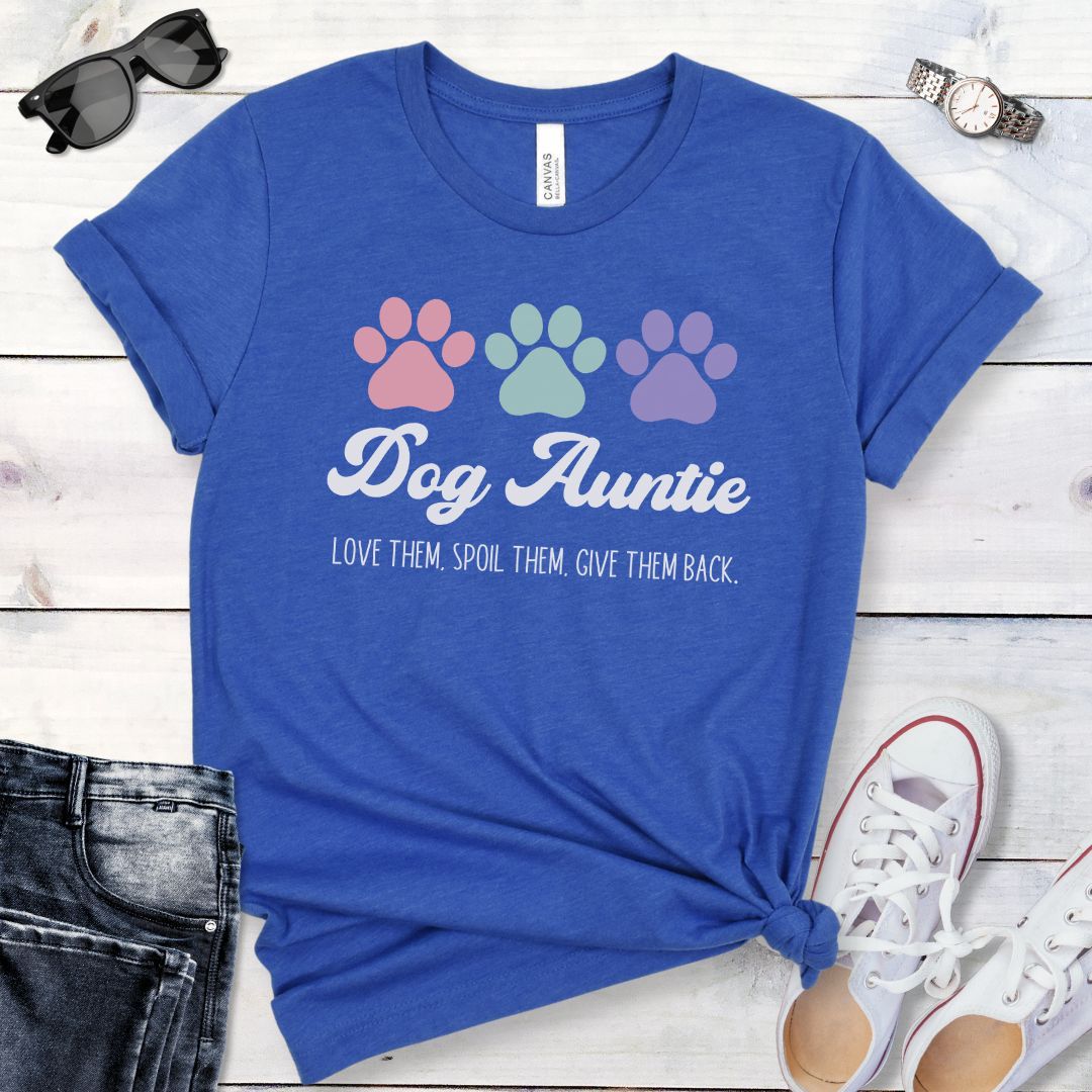 A royal blue dog lover t-shirt with 'Dog Auntie, Love Them Spoil Them, Give Theme Back' text and colorful paw prints design, placed on a wooden surface with jeans and white shoes in the background.