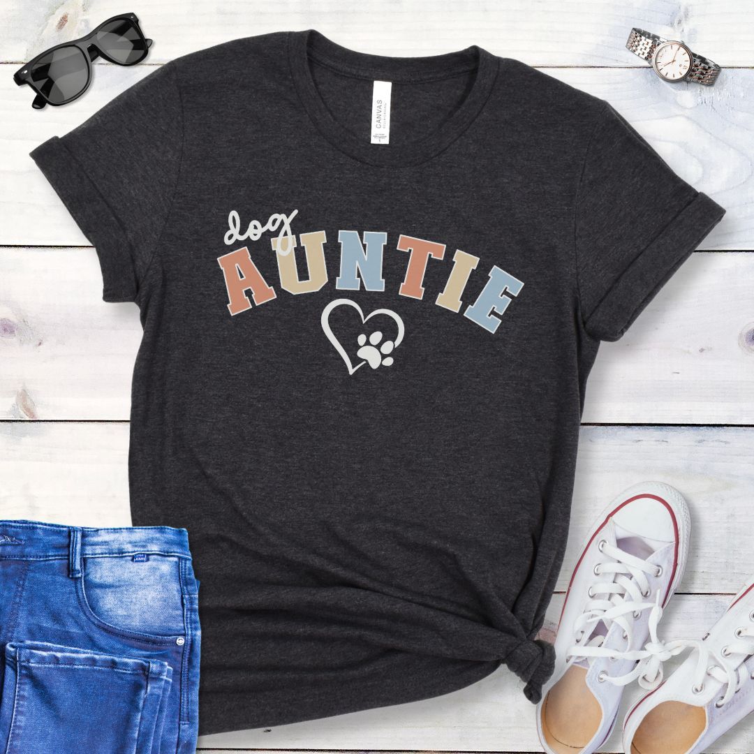 A dark grey dog lover t-shirt with 'Dog Auntie' text in multicolor, varsity style lettering and a heart and paw graphic, displayed with blue jeans, white shoes, and accessories.