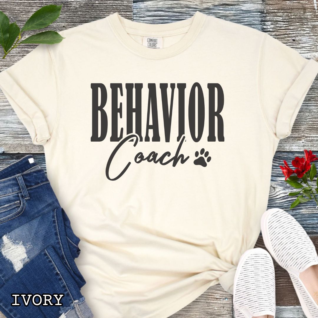 Beige (Ivory) Comfort Colors dog trainer t-shirt with 'BEHAVIOR Coach' text and small paw print, presented on a wooden surface with accessories.