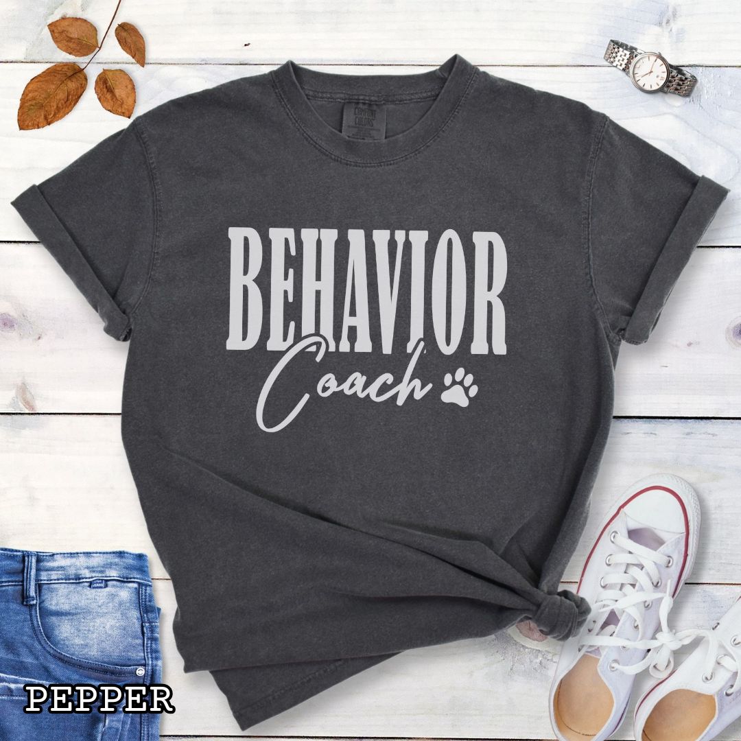 Dark Grey (Pepper) Comfort Colors dog trainer t-shirt with 'BEHAVIOR Coach' text and small paw print, presented on a wooden surface with accessories.
