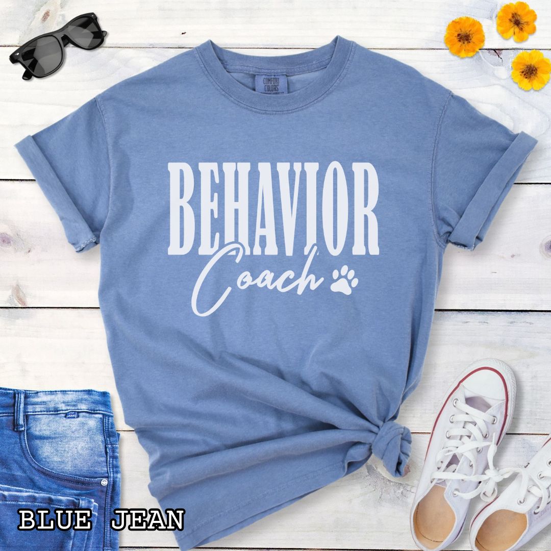 Blue Comfort Colors dog trainer t-shirt with 'BEHAVIOR Coach' text and small paw print, presented on a wooden surface with accessories.