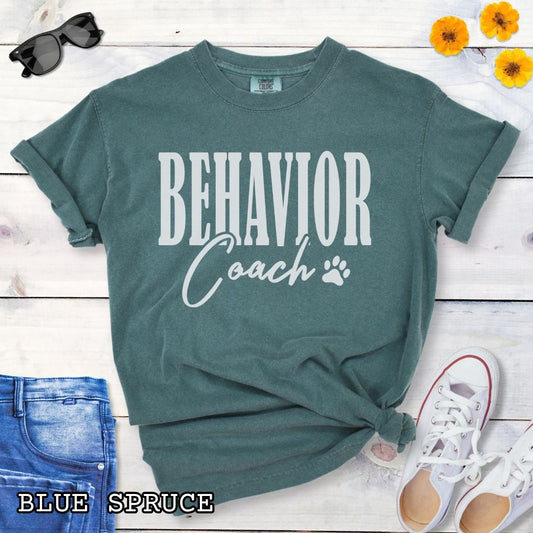 Green (Blue Spruce) Comfort Colors dog trainer t-shirt with 'BEHAVIOR Coach' text and small paw print, presented on a wooden surface with accessories.