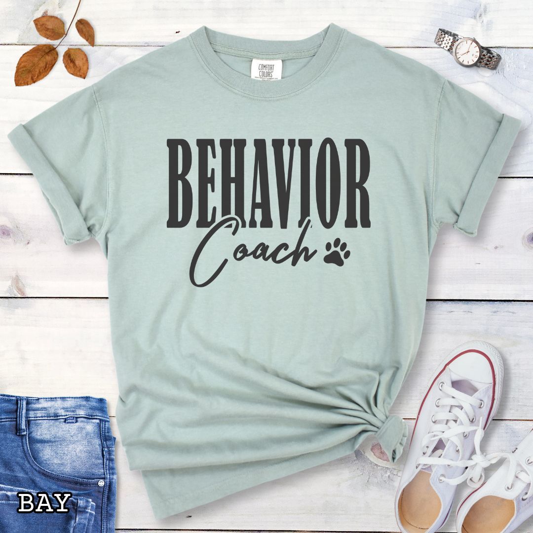 Light green (Bay) Comfort Colors dog trainer t-shirt with 'BEHAVIOR Coach' text and small paw print, presented on a wooden surface with accessories.