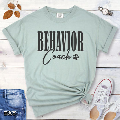 Light green (Bay) Comfort Colors dog trainer t-shirt with 'BEHAVIOR Coach' text and small paw print, presented on a wooden surface with accessories.