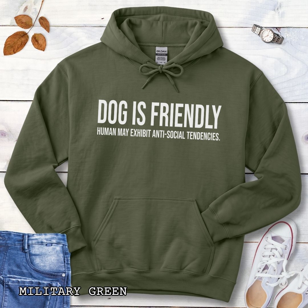 A military green pullover dog lover hoodie for introverts with bold, humorous text 'DOG IS FRIENDLY, HUMAN MAY EXHIBIT ANTI-SOCIAL TENDENCIES.' laid flat on a white wood background with decorative props including jeans, a watch, leaves, and sneakers.