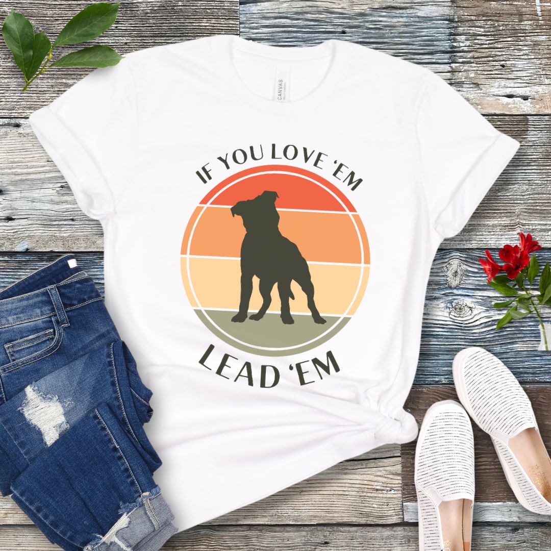 White dog training t-shirt with vintage sunset, dog graphic and text If You Love Them, Lead Them on a wooden surface with jeans and shoes.