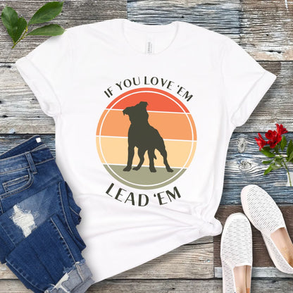 White dog training t-shirt with vintage sunset, dog graphic and text If You Love Them, Lead Them on a wooden surface with jeans and shoes.