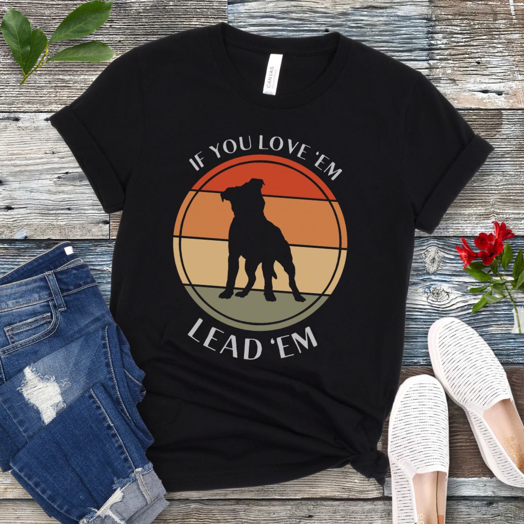 Black dog training t-shirt with vintage sunset, dog graphic and text If You Love Them, Lead Them on a wooden surface with jeans and shoes.