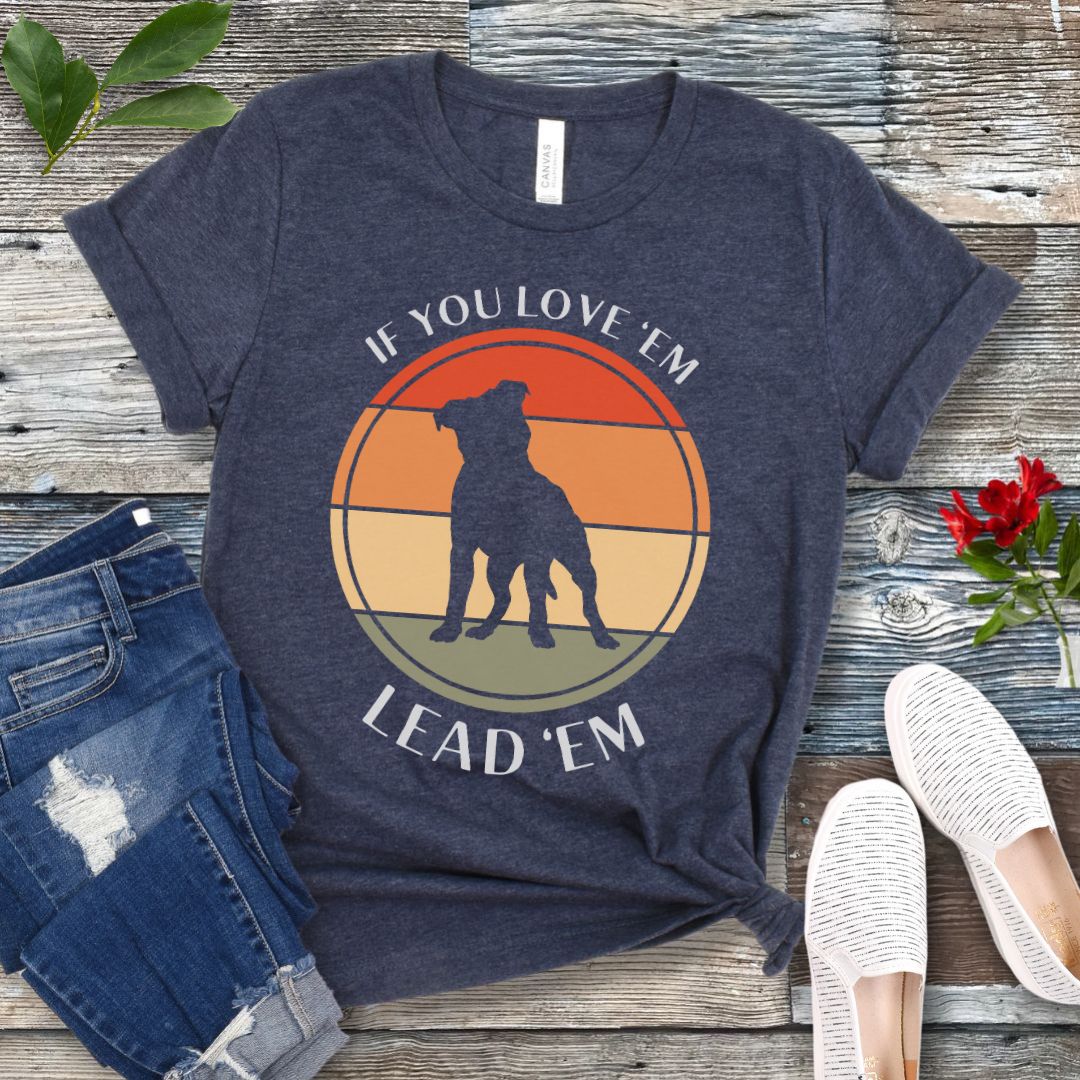 Heather navy blue dog training t-shirt with vintage sunset, dog graphic and text If You Love Them, Lead Them on a wooden surface with jeans and shoes.
