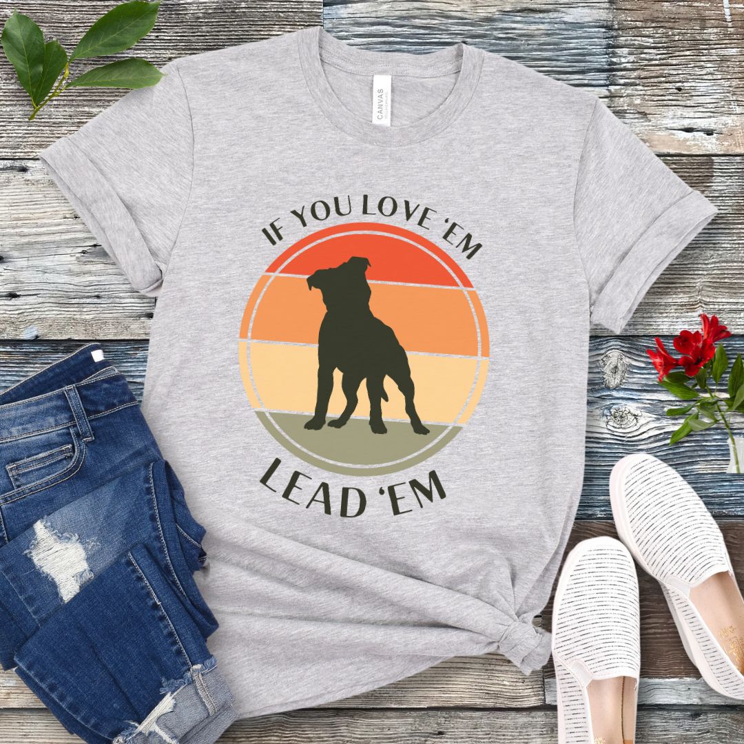 Light grey dog training t-shirt with vintage sunset, dog graphic and text If You Love Them, Lead Them on a wooden surface with jeans and shoes.