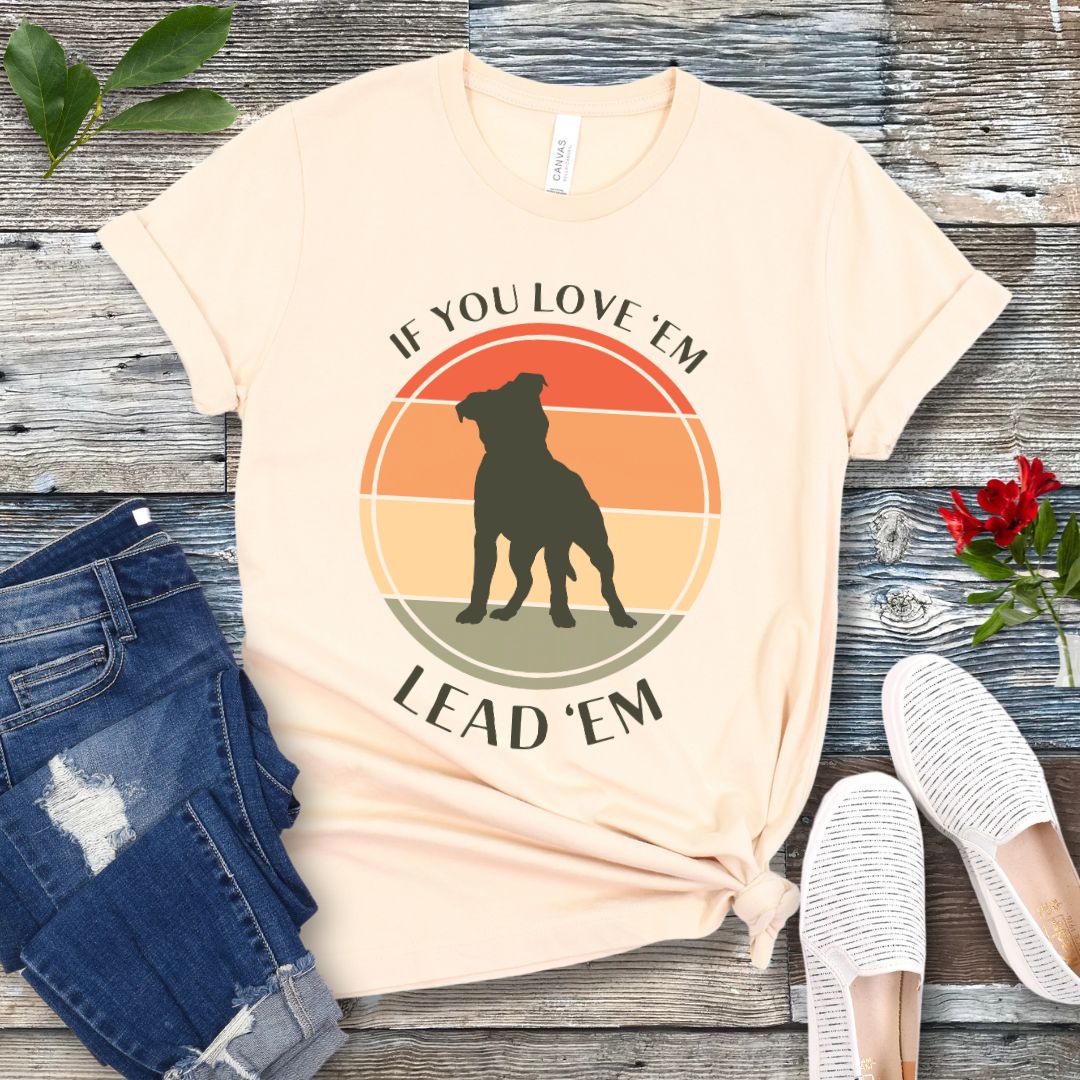 Beige dog training t-shirt with vintage sunset, dog graphic and text If You Love Them, Lead Them on a wooden surface with jeans and shoes.