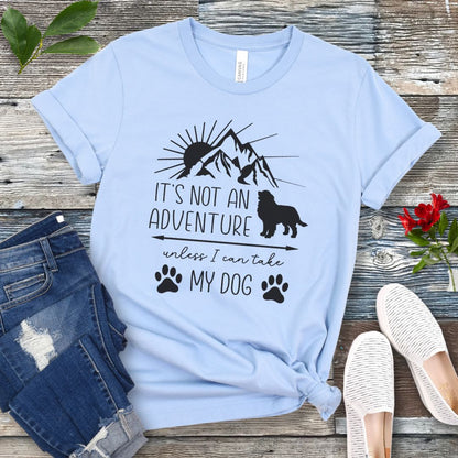 A baby blue dog lover T-shirt with a graphic that says 'IT'S NOT AN ADVENTURE UNLESS I CAN TAKE MY DOG' accompanied by images of a mountain, a dog and paw prints. The shirt is displayed alongside a pair of jeans and a pair of white shoes on a wooden background.