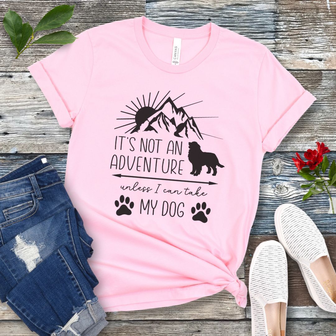 A pink dog lover T-shirt with a graphic that says 'IT'S NOT AN ADVENTURE UNLESS I CAN TAKE MY DOG' accompanied by images of a mountain, a dog and paw prints. The shirt is displayed alongside a pair of jeans and a pair of white shoes on a wooden background.