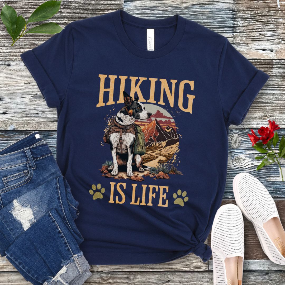 Navy blue t-shirt with an image of a dog on a mountain trial and text 'HIKING IS LIFE', displayed on a wooden surface with a pair of jeans and white shoes in the background.