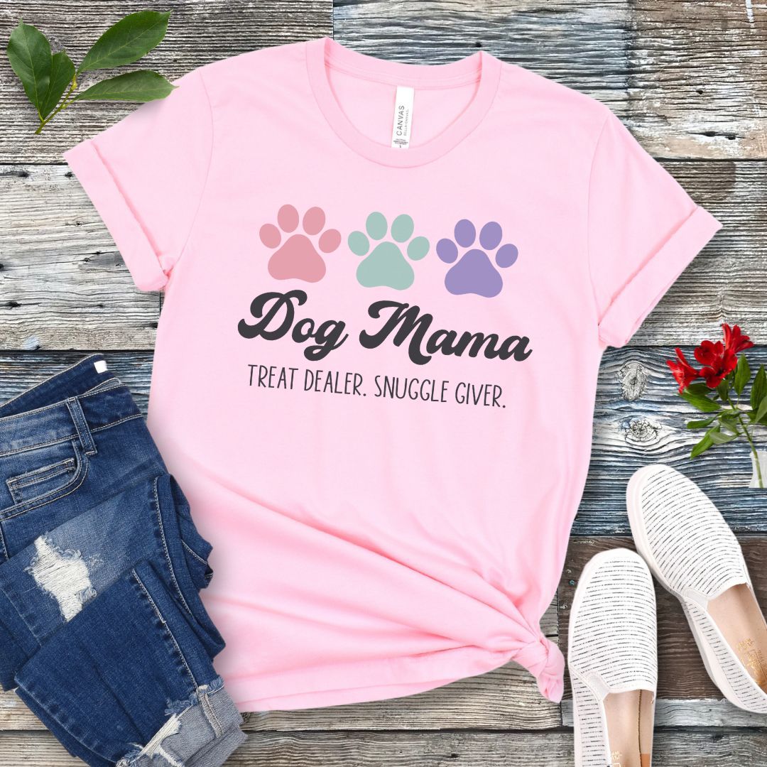 A pink dog lover t-shirt with 'Dog Mama, Treat Dealer, Snuggle Giver' text and pastel paw prints design, displayed with jeans and white shoes on a wooden background.