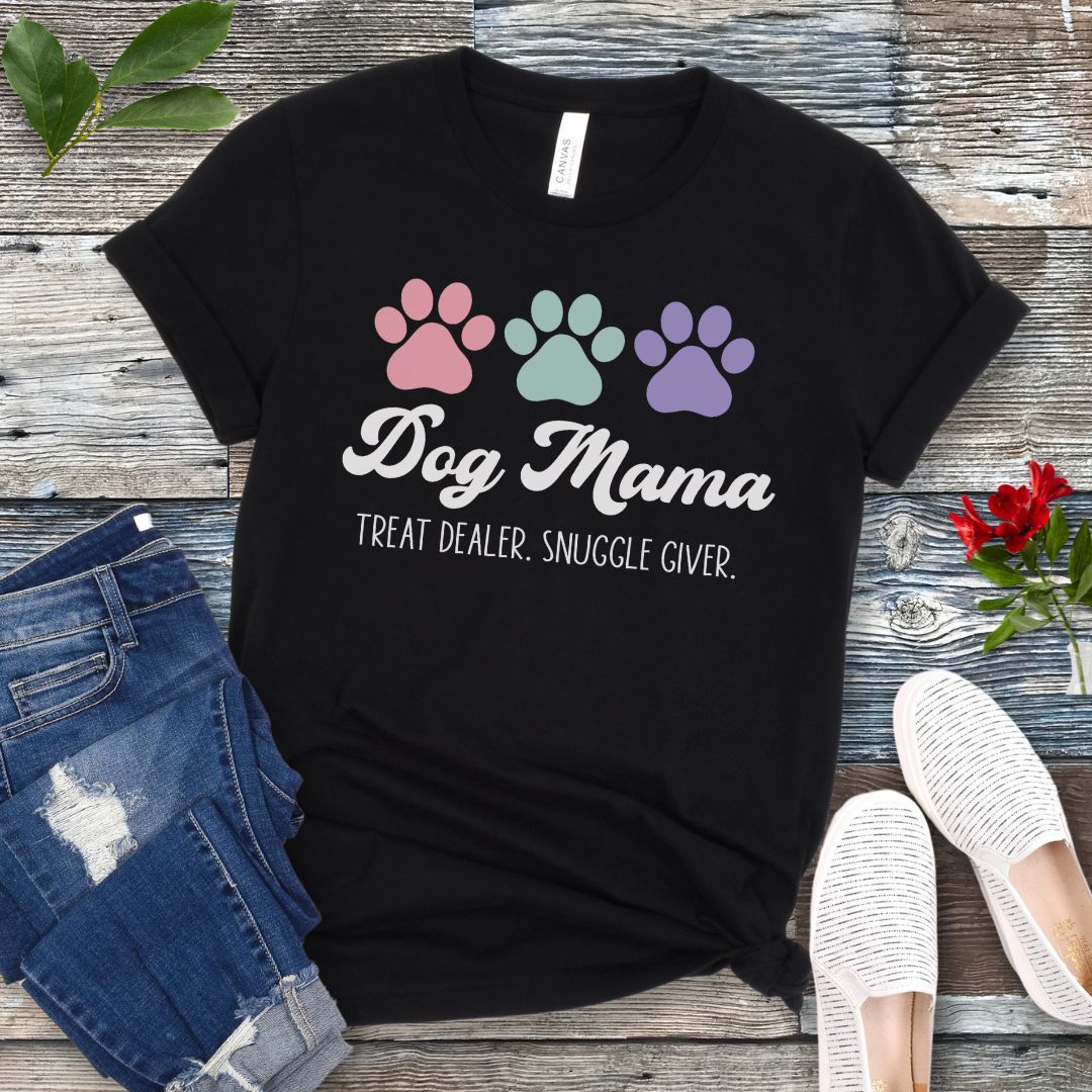 A black dog lover t-shirt with 'Dog Mama, Treat Dealer, Snuggle Giver' text and pastel paw prints design, displayed with jeans and white shoes on a wooden background.