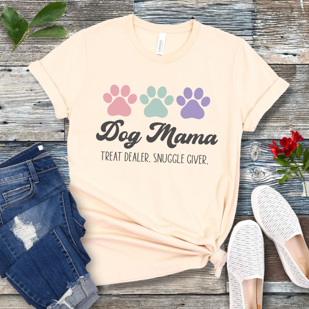 A beige dog lover t-shirt with 'Dog Mama, Treat Dealer, Snuggle Giver' text and pastel paw prints design, displayed with jeans and white shoes on a wooden background.