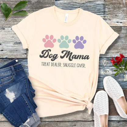 A beige dog lover t-shirt with 'Dog Mama, Treat Dealer, Snuggle Giver' text and pastel paw prints design, displayed with jeans and white shoes on a wooden background.