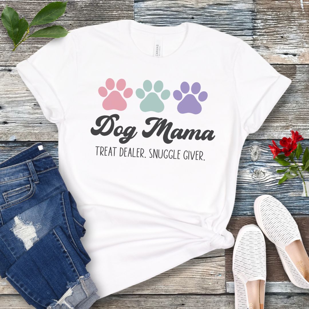 A white dog lover t-shirt with 'Dog Mama, Treat Dealer, Snuggle Giver' text and pastel paw prints design, displayed with jeans and white shoes on a wooden background.