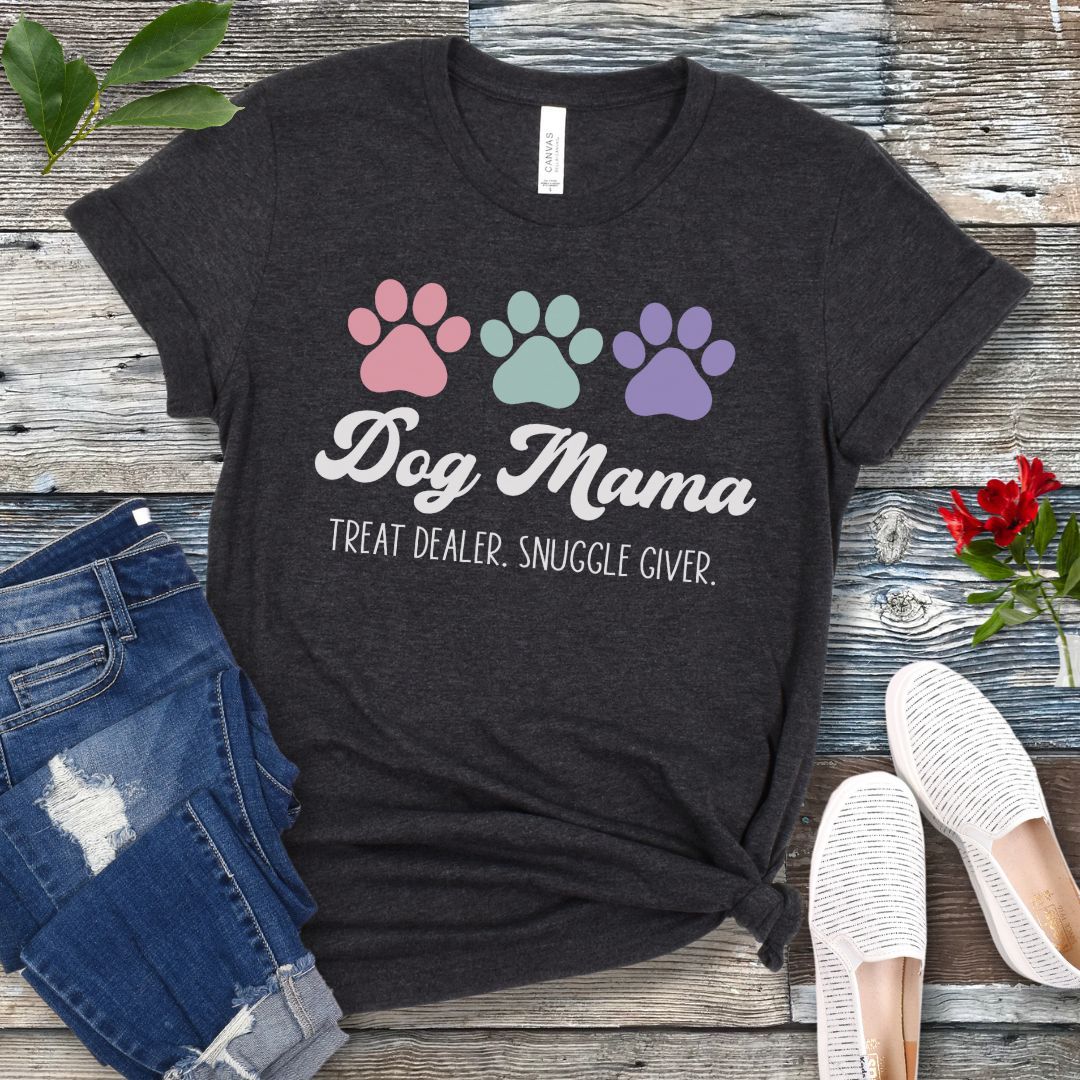 A dark grey dog lover t-shirt with 'Dog Mama, Treat Dealer, Snuggle Giver' text and pastel paw prints design, displayed with jeans and white shoes on a wooden background.