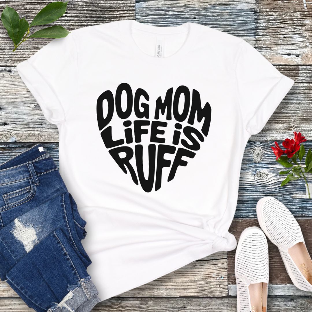 A white t-shirt for dog lovers with the text 'DOG MOM LIFE IS RUFF' printed in a heart shape, placed on a wooden surface with a pair of blue jeans and white shoes in the background.