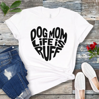 A white t-shirt for dog lovers with the text 'DOG MOM LIFE IS RUFF' printed in a heart shape, placed on a wooden surface with a pair of blue jeans and white shoes in the background.