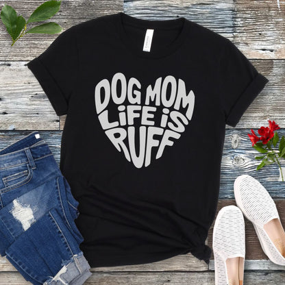 A black t-shirt for dog lovers with the text 'DOG MOM LIFE IS RUFF' printed in a heart shape, placed on a wooden surface with a pair of blue jeans and white shoes in the background.