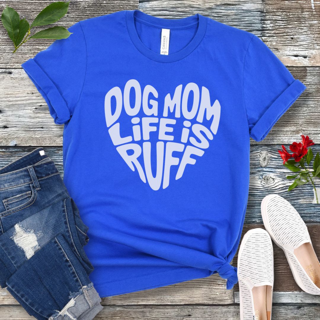 A royal blue t-shirt for dog lovers with the text 'DOG MOM LIFE IS RUFF' printed in a heart shape, placed on a wooden surface with a pair of blue jeans and white shoes in the background.