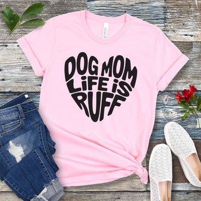 A pink t-shirt for dog lovers with the text 'DOG MOM LIFE IS RUFF' printed in a heart shape, placed on a wooden surface with a pair of blue jeans and white shoes in the background.