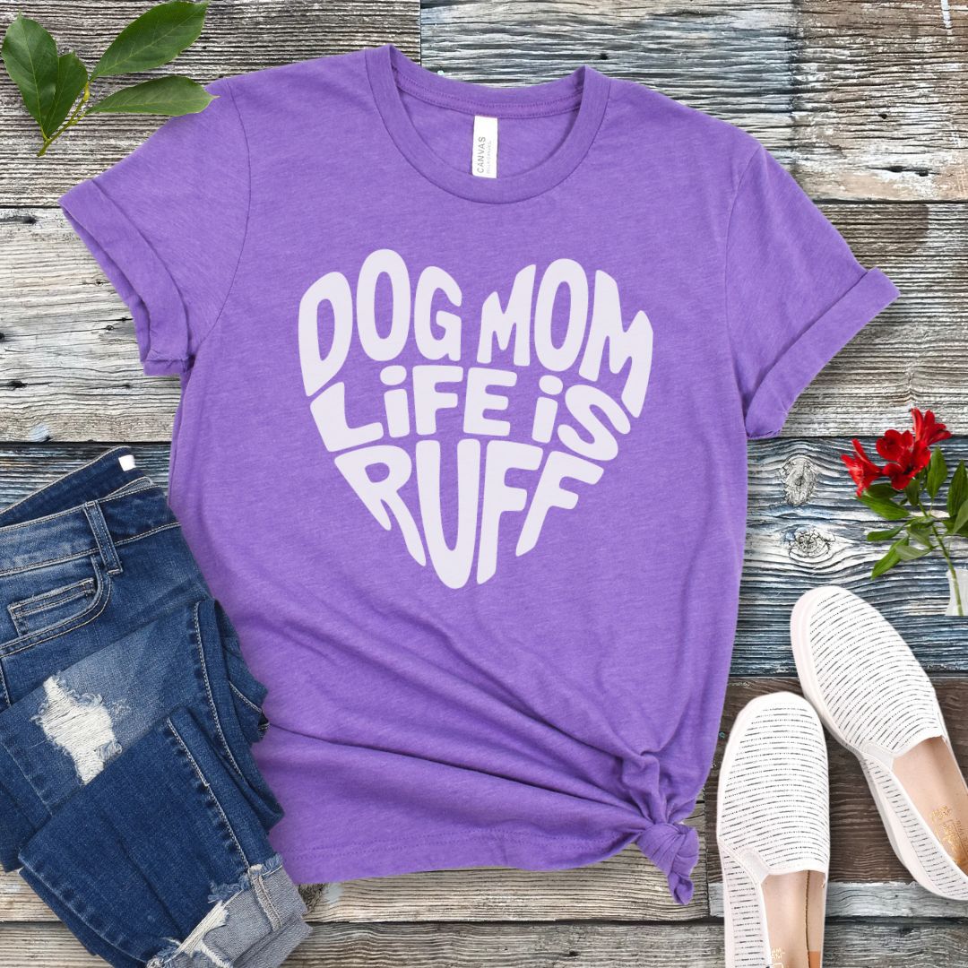 A purple t-shirt for dog lovers with the text 'DOG MOM LIFE IS RUFF' printed in a heart shape, placed on a wooden surface with a pair of blue jeans and white shoes in the background.