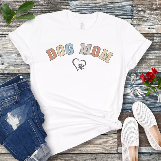 A white dog lover t-shirt with 'DOG MOM' written on it in bold, multicolor, varsity-style letters, with a heart and paw print underneath the text, displayed on a wooden background alongside jeans, white shoes, and a red flower.