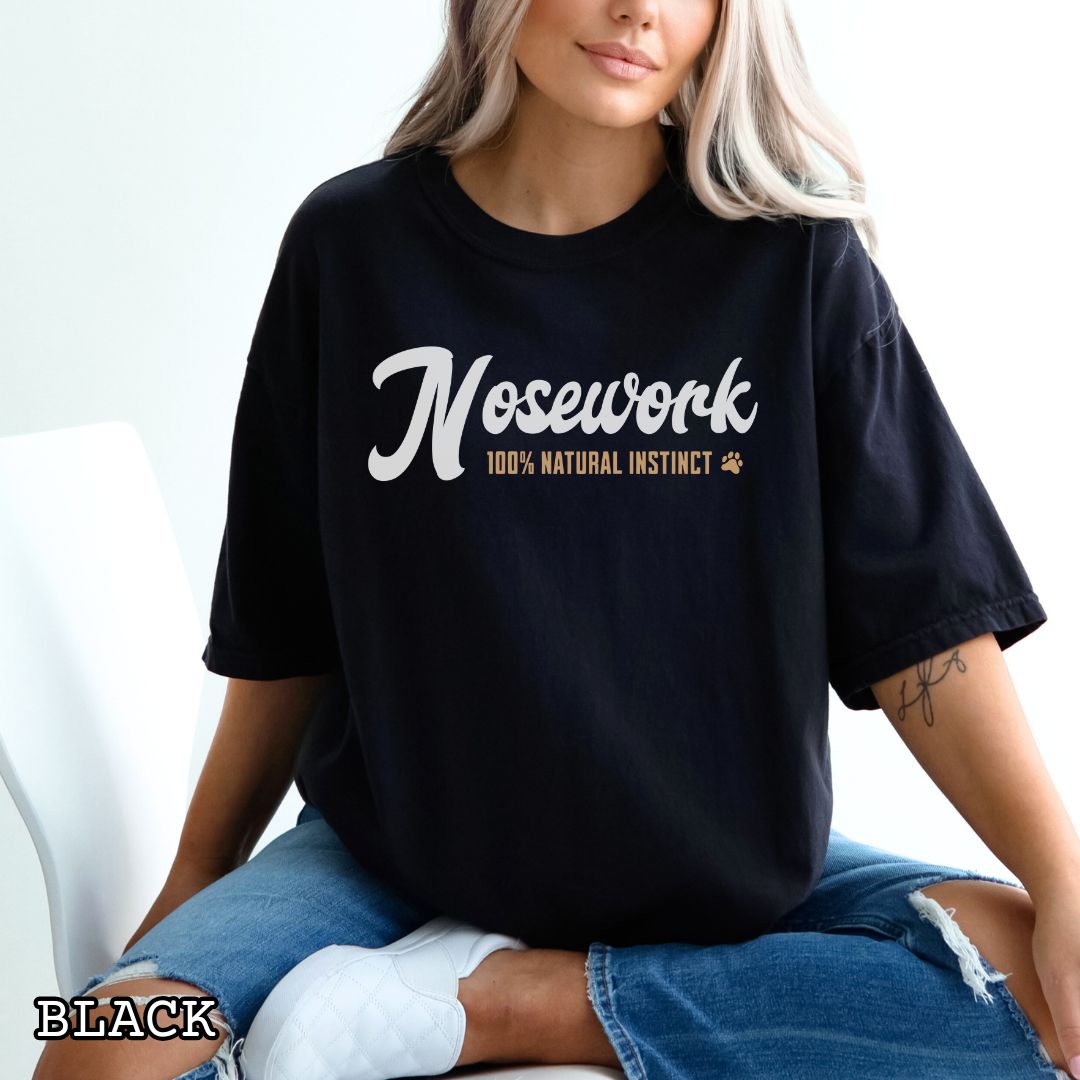 Person wearing a black Comfort Colors t-shirt with 'Nosework 100% Natural Instinct' text, sitting on a white couch.