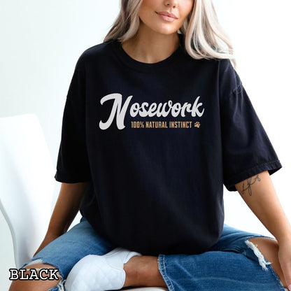 Person wearing a black Comfort Colors t-shirt with 'Nosework 100% Natural Instinct' text, sitting on a white couch.
