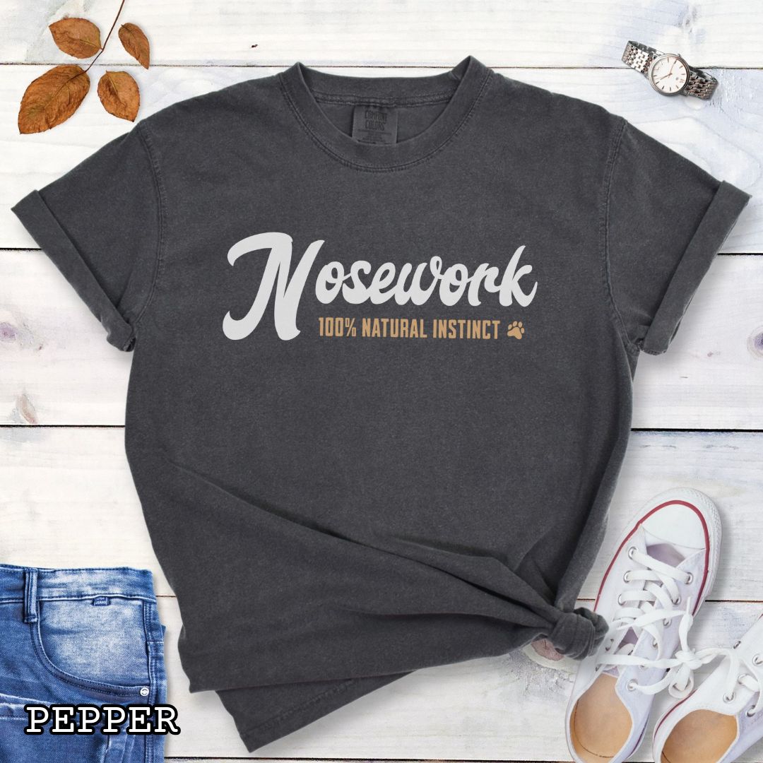 Grey (Pepper) Comfort Colors dog sports t-shirt with 'Nosework 100% Natural Instinct' text on a wooden surface with jeans and sneakers.