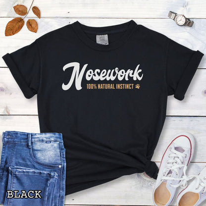 Black Comfort Colors dog sports t-shirt with 'Nosework 100% Natural Instinct' text on a wooden surface with jeans and sneakers.