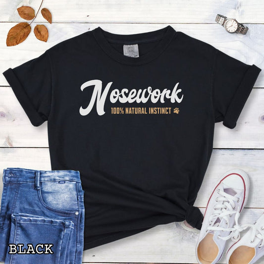 Black Comfort Colors dog sports t-shirt with 'Nosework 100% Natural Instinct' text on a wooden surface with jeans and sneakers.