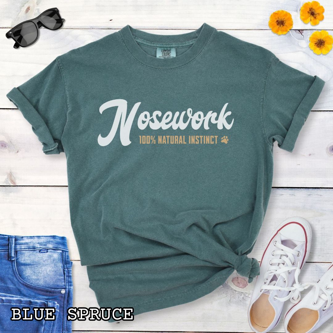 Green (Blue Spruce) Comfort Colors dog sports t-shirt with 'Nosework 100% Natural Instinct' text on a wooden surface with jeans and sneakers.