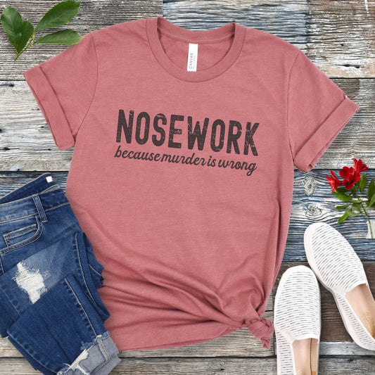 Mauve dog sports t-shirt with 'Nosework because murder is wrong' text on a wooden surface with jeans and shoes.