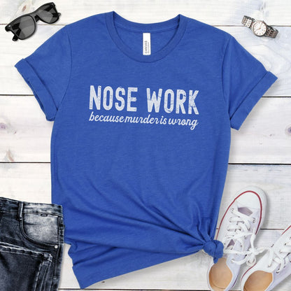 Blue t-shirt for dog scent work trainers with 'Nosework because murder is wrong' humorous text on a wooden surface with jeans and shoes.