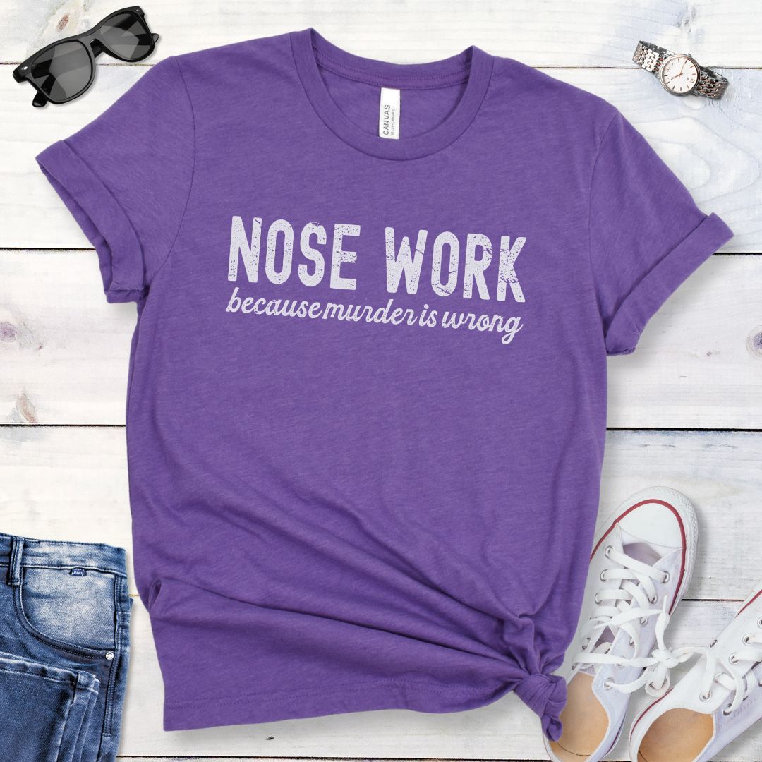 Purple t-shirt for dog scent work trainers with 'Nosework because murder is wrong' humorous text on a wooden surface with jeans and shoes.