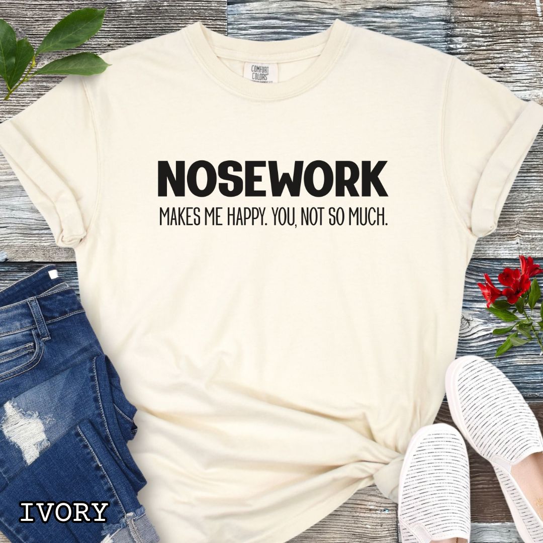 Beige Comfort Colors t-shirt with 'NOSEWORK MAKES ME HAPPY. YOU NOT SO MUCH.' text, shown on a wooden surface with denim jeans and a flower.