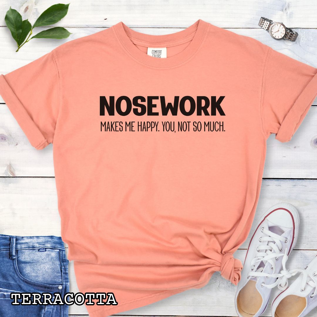 Orange Comfort Colors t-shirt with 'NOSEWORK MAKES ME HAPPY. YOU NOT SO MUCH.' text, shown on a wooden surface with denim jeans and a flower.