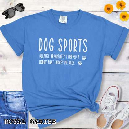Royal blue Comfort Colors t-shirt with humorous text 'Dog Sports, Because Apparently I Needed a Hobby That Judges Me Back' accompanied by paw prints, shown on a wooden surface with jeans and flowers.