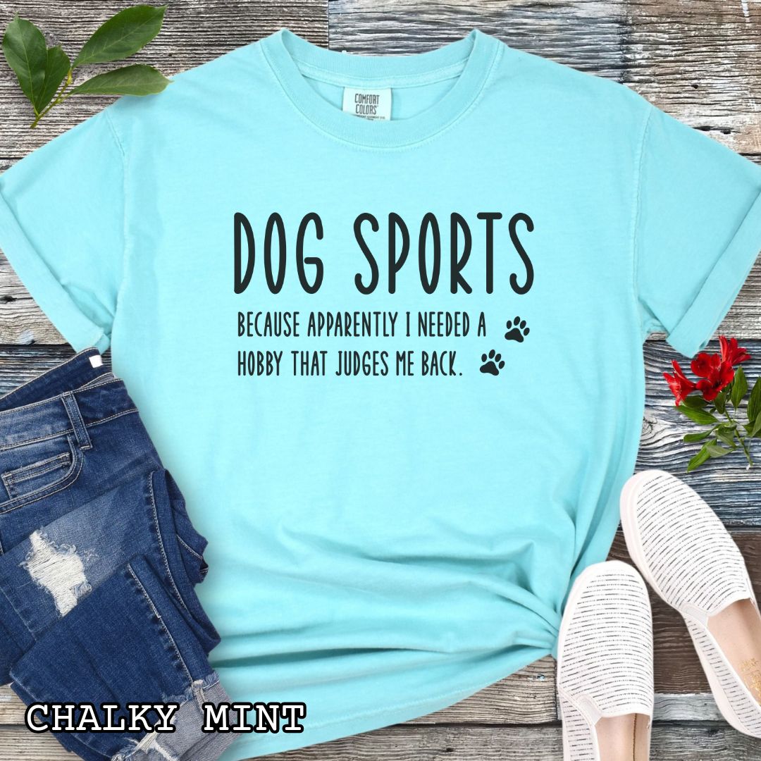 Mint green Comfort Colors t-shirt with humorous text 'Dog Sports, Because Apparently I Needed a Hobby That Judges Me Back' accompanied by paw prints, shown on a wooden surface with jeans and flowers.