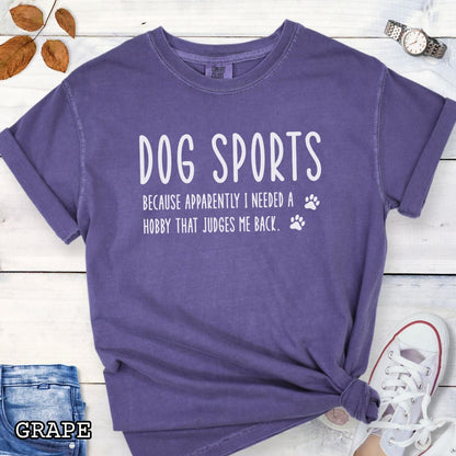 Purple Comfort Colors t-shirt with humorous text 'Dog Sports, Because Apparently I Needed a Hobby That Judges Me Back' accompanied by paw prints, shown on a wooden surface with jeans and flowers.