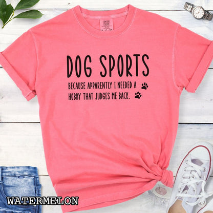Pink Comfort Colors t-shirt with humorous text 'Dog Sports, Because Apparently I Needed a Hobby That Judges Me Back' accompanied by paw prints, shown on a wooden surface with jeans and flowers.